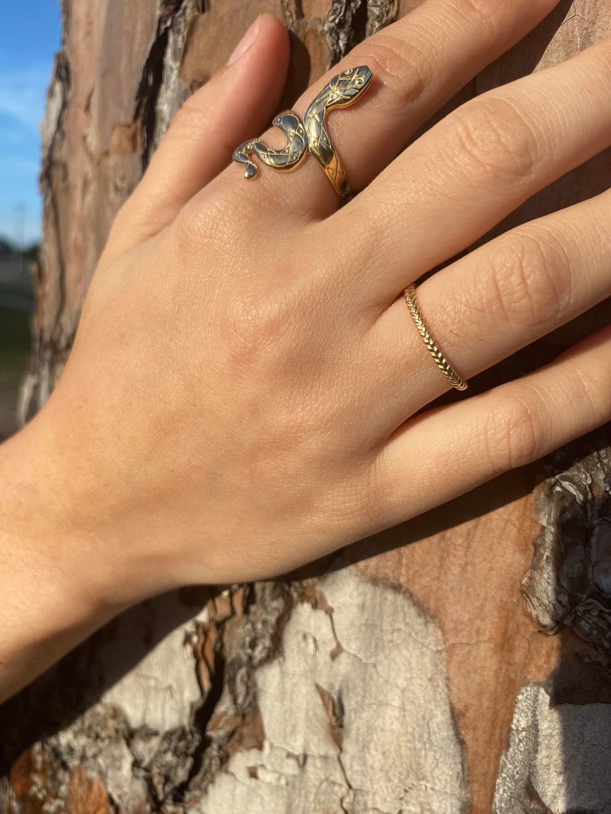 Hand wearing 14k gold slithering snake ring with intricate patterns wrapped around finger against a tree bark background